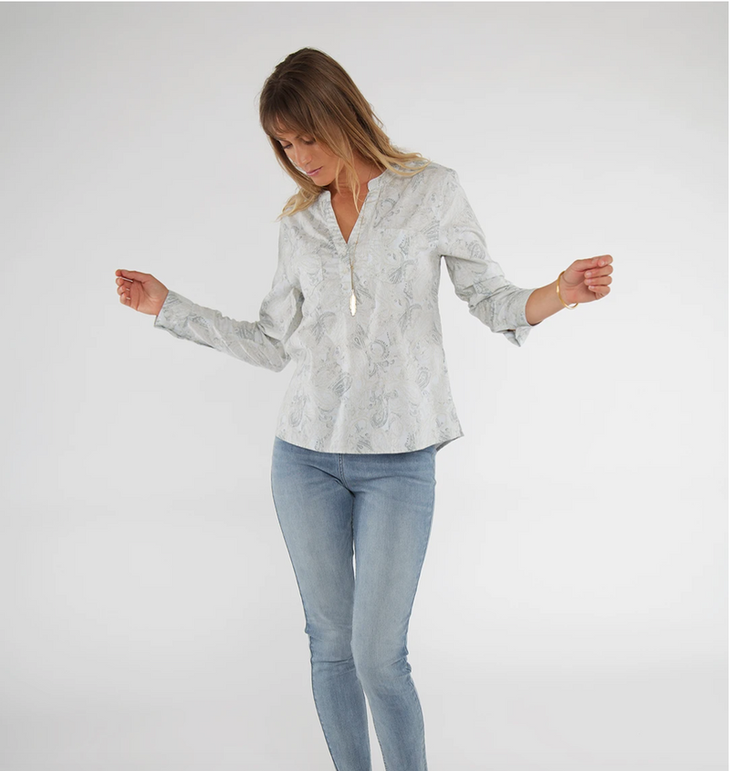This is a front view of the Carve Designs Dylan Twill shirt in Quarry Lily. The shirt is white with a paisley pattern in greys and blues. It has a mandarin collar with v-neck and a bottom button. It has 2 chest pockets and long sleeves. The shirt hits at hip level on the model.