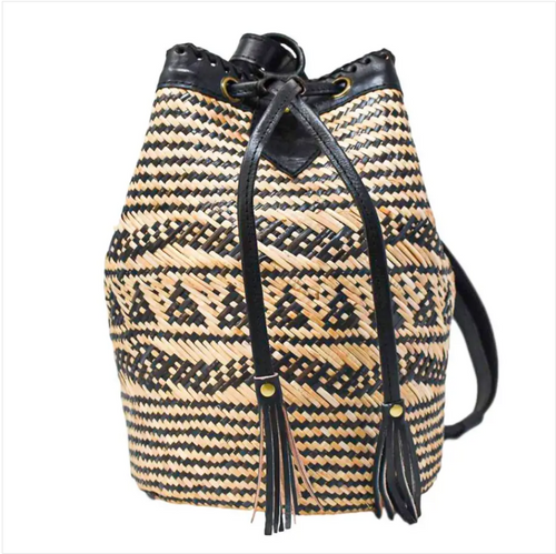 The Delilah Bucket bag is a backpack style, cinch top bag with a black and natural straw woven pattern and black leather straps, tassels and accents.