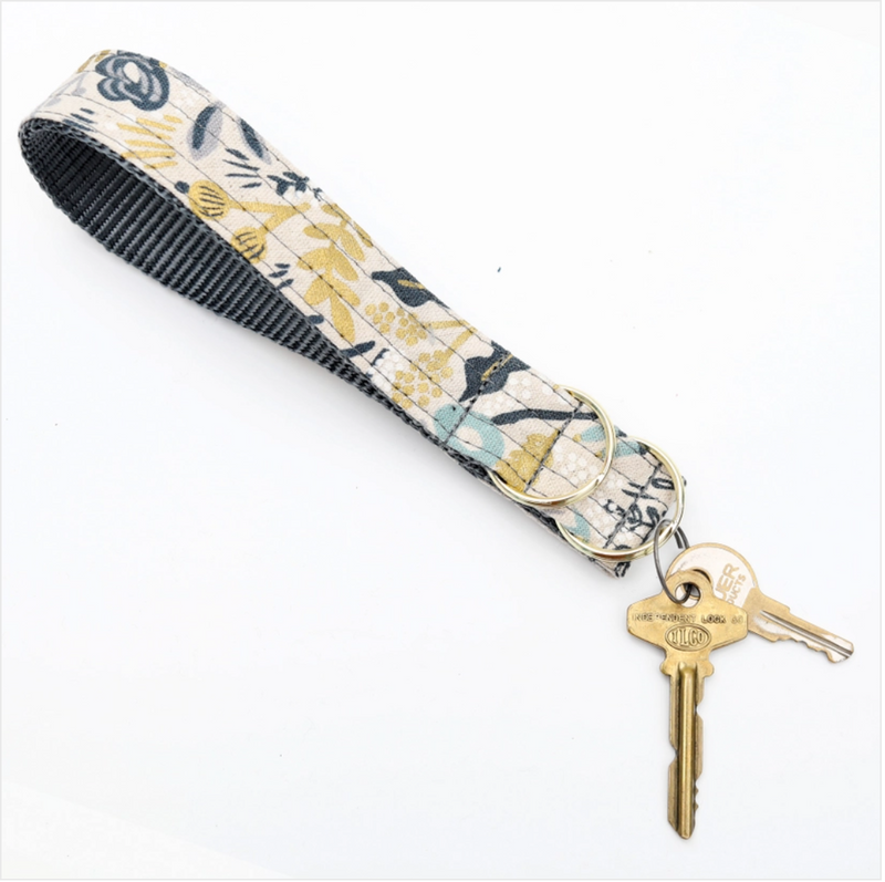 A blue and gold tone floral print key fob with a blue interior strap. There are two key rings for key attachment.