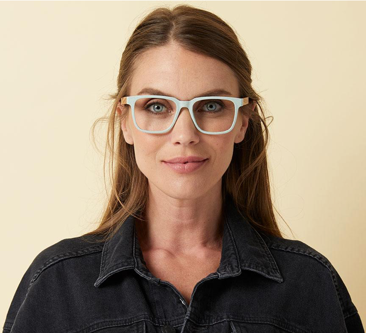 A woman modeling the Peepers Homespun reading glasses in mint/wood. These frames have a speckled, matte appearance with zebrawood temples.