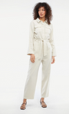 Model is wearing the Ludlow Jumpsuit, in oatmeal. It is a long sleeve, slightly cropped pant jumpsuit with a button front, two chest pockets and tie fabric belt.