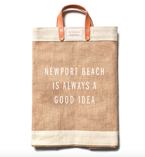 The Apolis Market bag in natural tan, with a light cream colored top/bottom band and natural tone leather handles. The text NEWPORT BEACH IS ALWAYS A GOOOD IDEA is printed on the front in white.