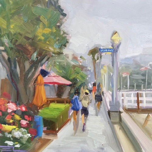 walkers on the walkway surrounding Balboa Island.  Houses on the left.  Trees, hedges, flowers and a red umbrella.  On the right docks to the bay with American Flags.  Cross street sign is Sapphire.